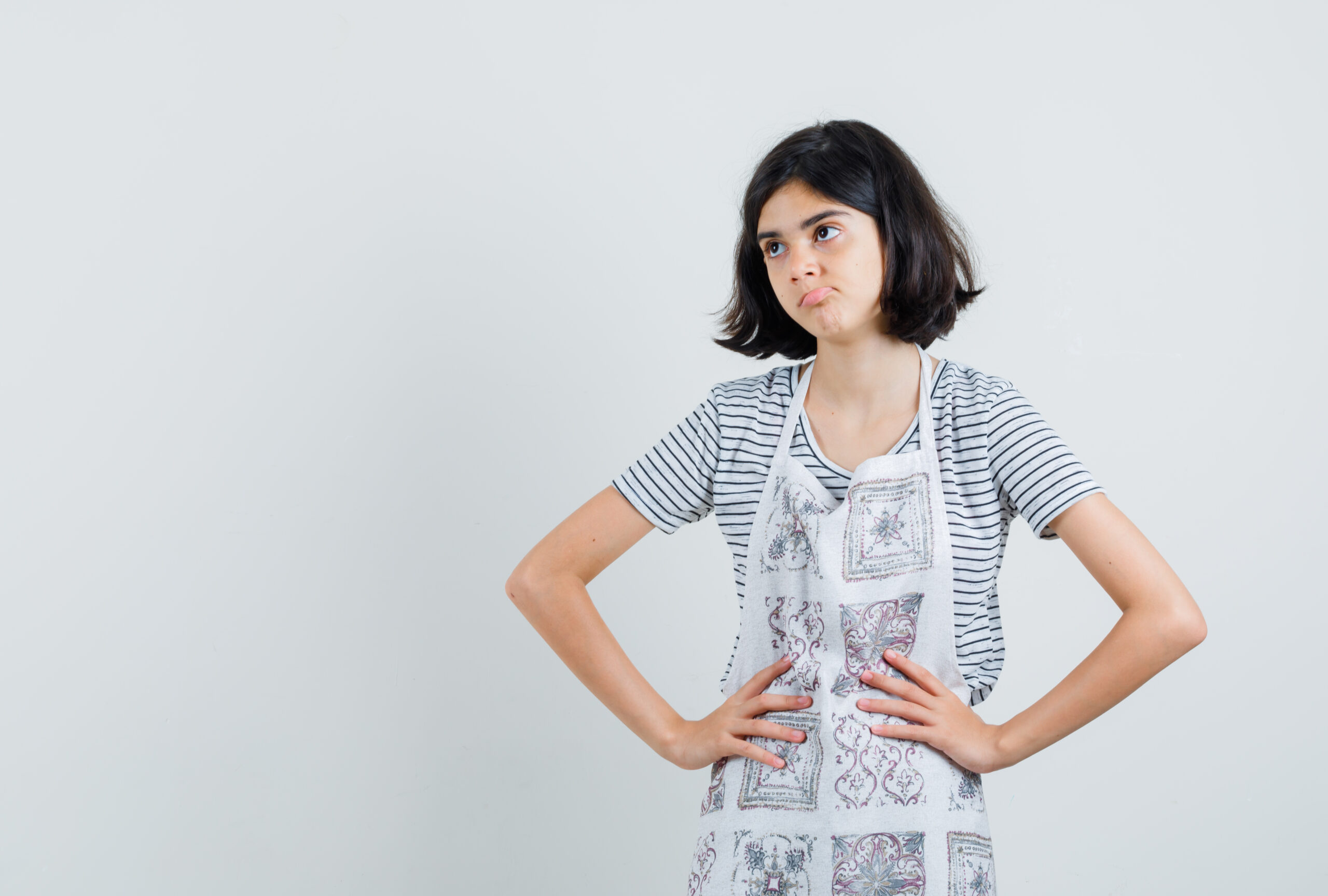 Little girl holding hands on waist in t-shirt, apron and looking pensive. front view.